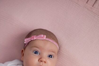 girl baby infant with a bow lies on the bed in a white bodysuit with a doll toy next to it, the child has a pink number 2 months on a pink background.